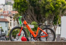 Bicicletas elétricas estacionadas ao ar livre, com árvores ao fundo.