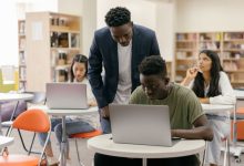 Estudantes participam de uma atividade em sala de aula enquanto uma professora os orienta; há laptops sobre as mesas.