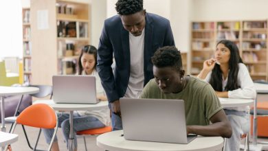 Estudantes participam de uma atividade em sala de aula enquanto uma professora os orienta; há laptops sobre as mesas.