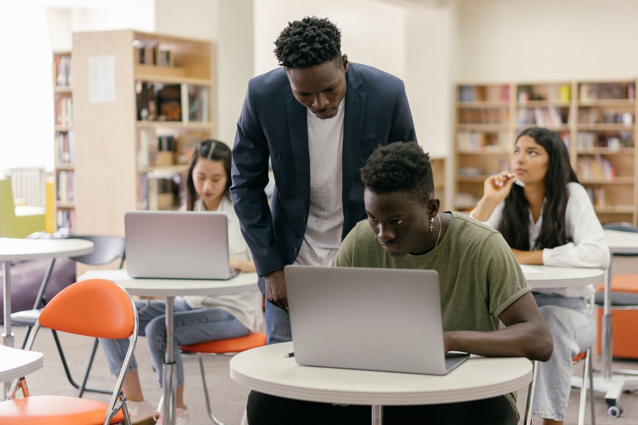 Estudantes participam de uma atividade em sala de aula enquanto uma professora os orienta; há laptops sobre as mesas.